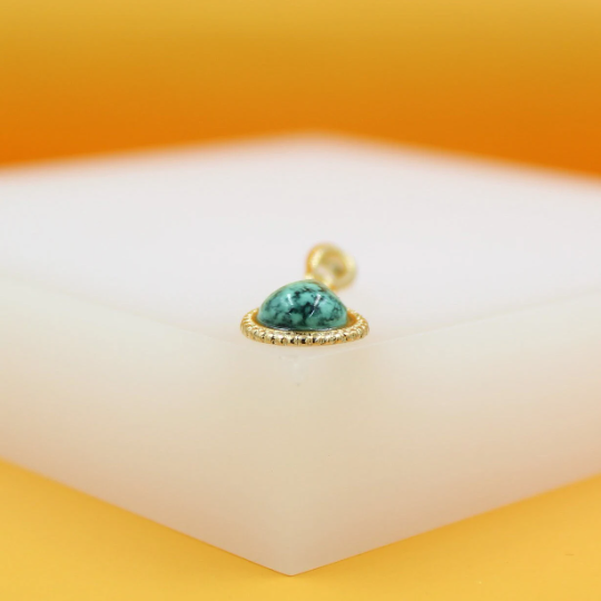 A gold pendant features a smooth, domed turquoise stone set on a white display surface against a warm yellow background.