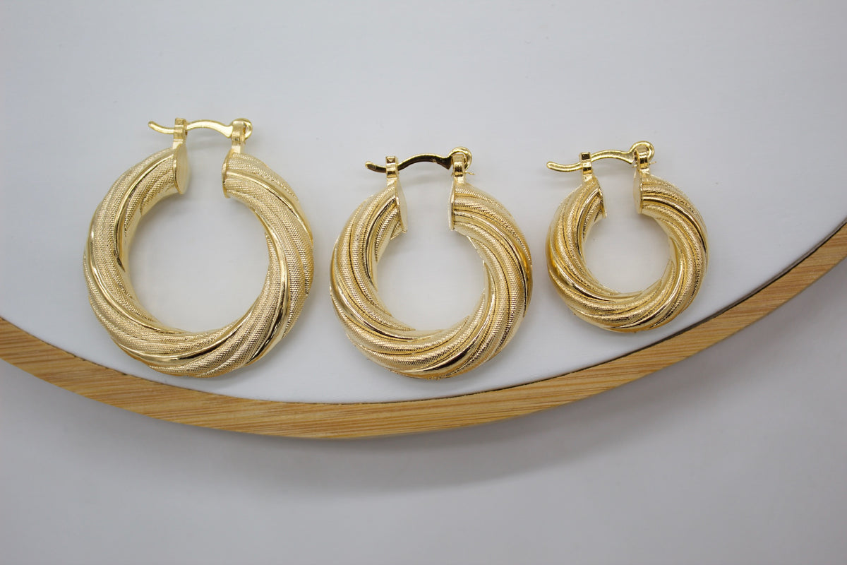 Three graduated sizes of textured gold hoop earrings rest on a white surface within a wooden display tray.