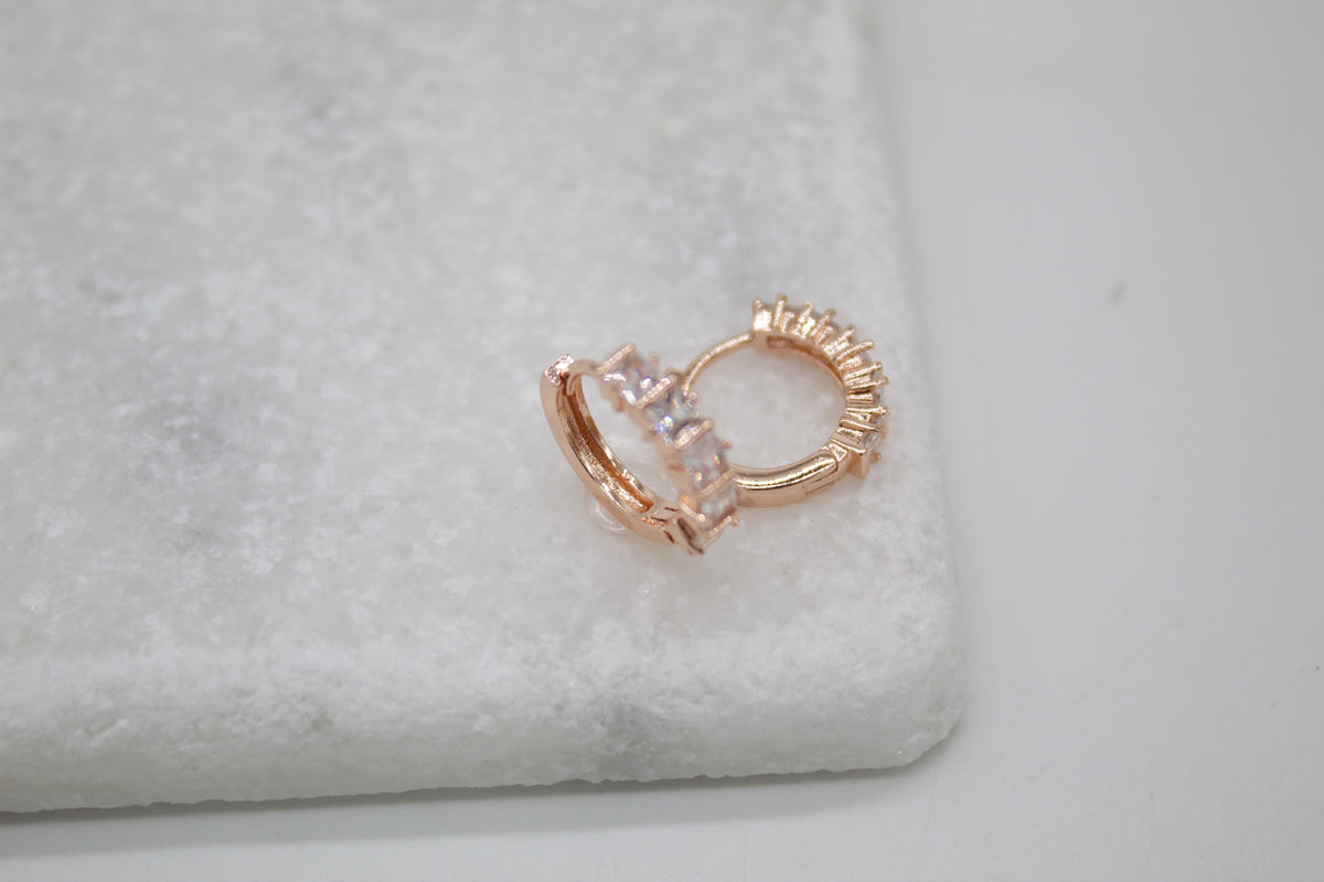 A pair of rose gold huggie hoop earrings with sparkling cubic zirconia stones rests on a textured white marble surface.