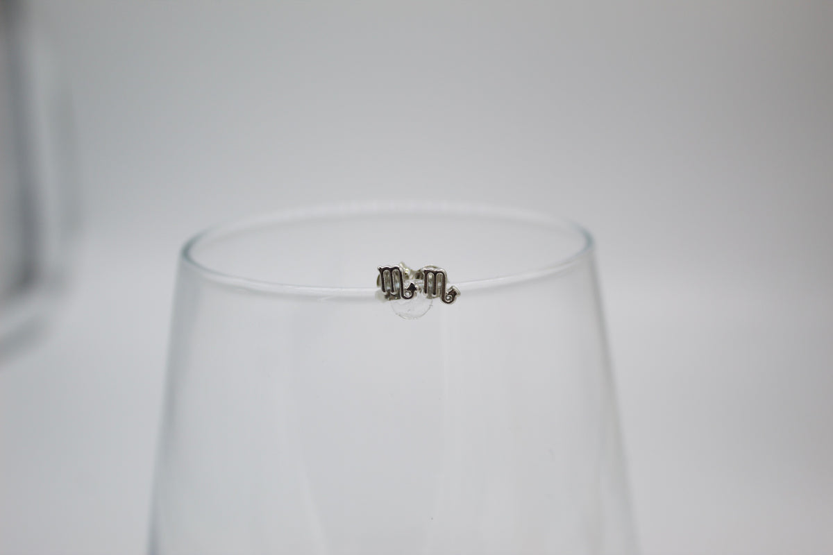 A pair of silver Scorpio zodiac sign stud earrings rests delicately on the rim of a clear glass.