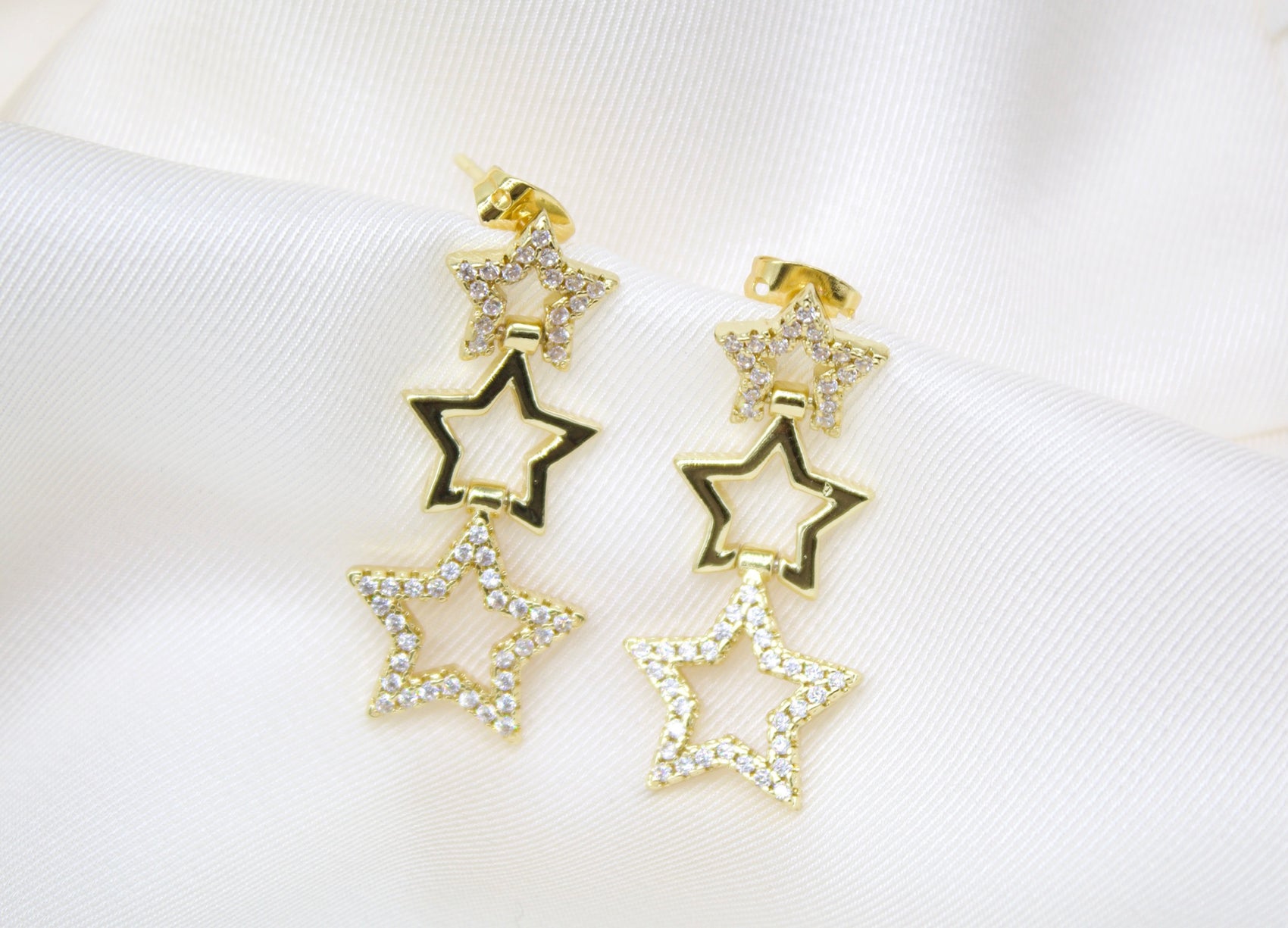 A pair of dangling gold star earrings sparkle against a soft white fabric background.