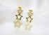 A pair of dangling gold star earrings sparkle against a soft white fabric background.
