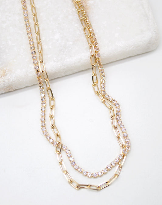 A layered gold chain necklace features a delicate rhinestone tennis strand and a chunky paperclip link chain resting against a white surface.