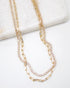 A layered gold necklace features a sparkling rhinestone tennis chain and a delicate paperclip chain resting on a white marble surface.
