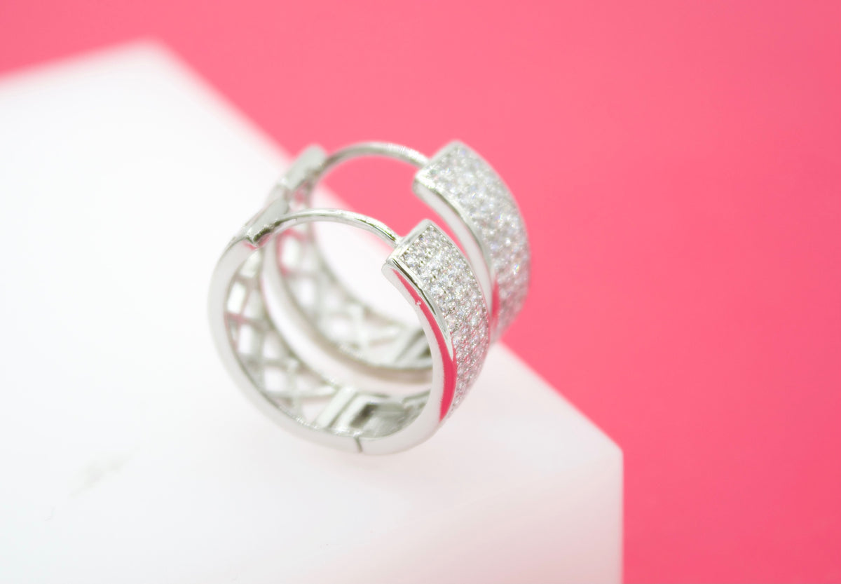 A pair of sparkling pave huggie hoop earrings rests on a white display against a vibrant pink background.
