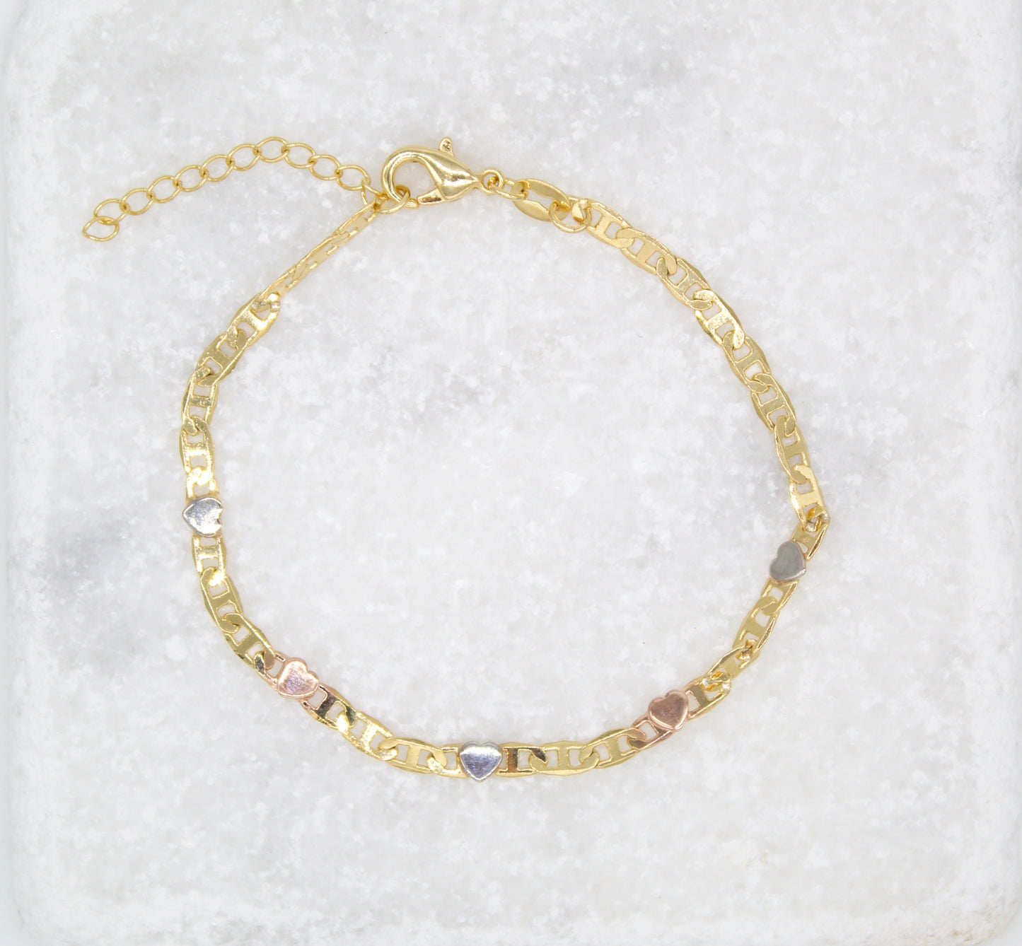 A delicate gold chain bracelet features alternating multi-toned heart charms and rests on a textured white marble surface.