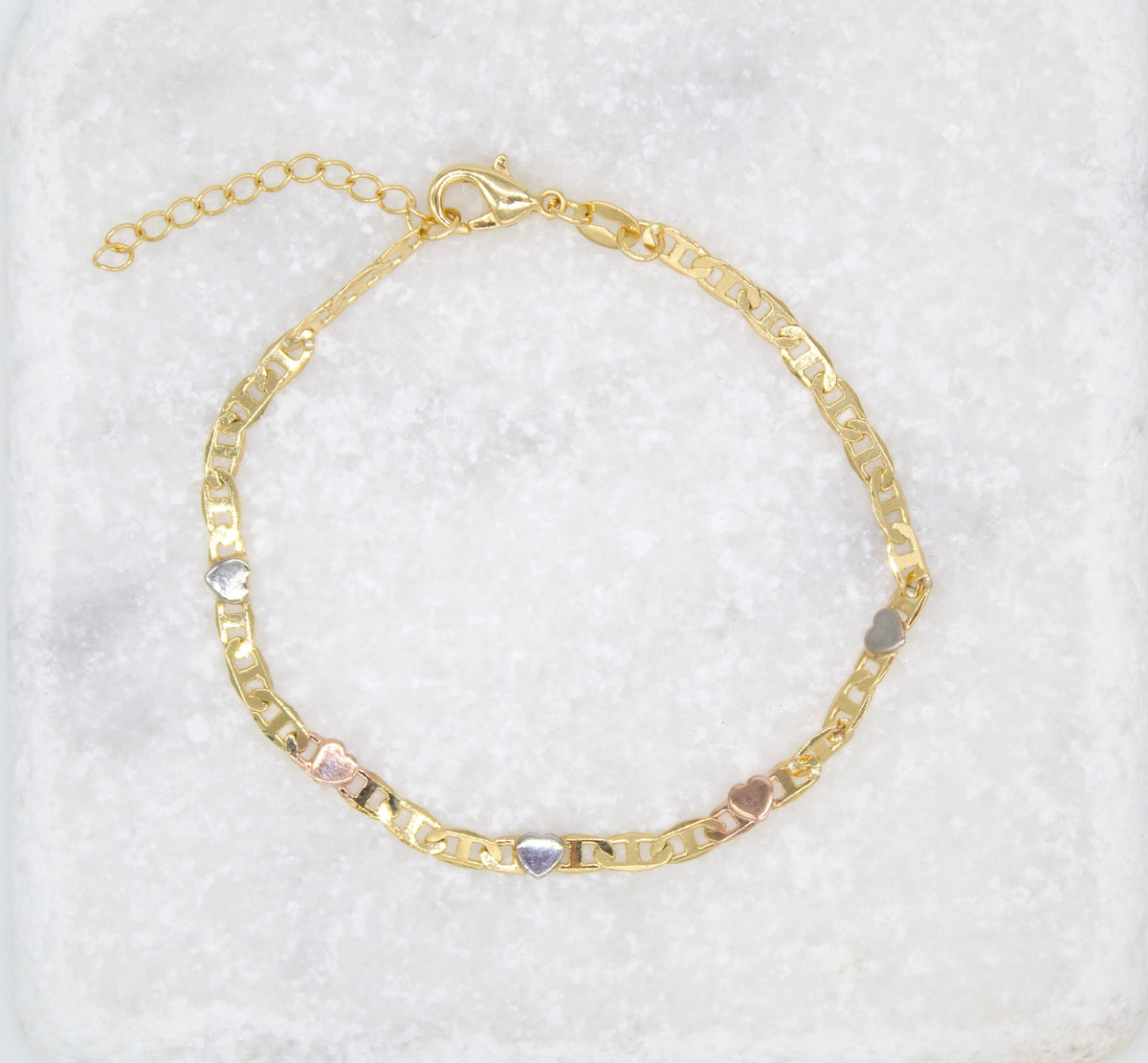 A delicate gold chain bracelet features alternating multi-toned heart charms and rests on a textured white marble surface.