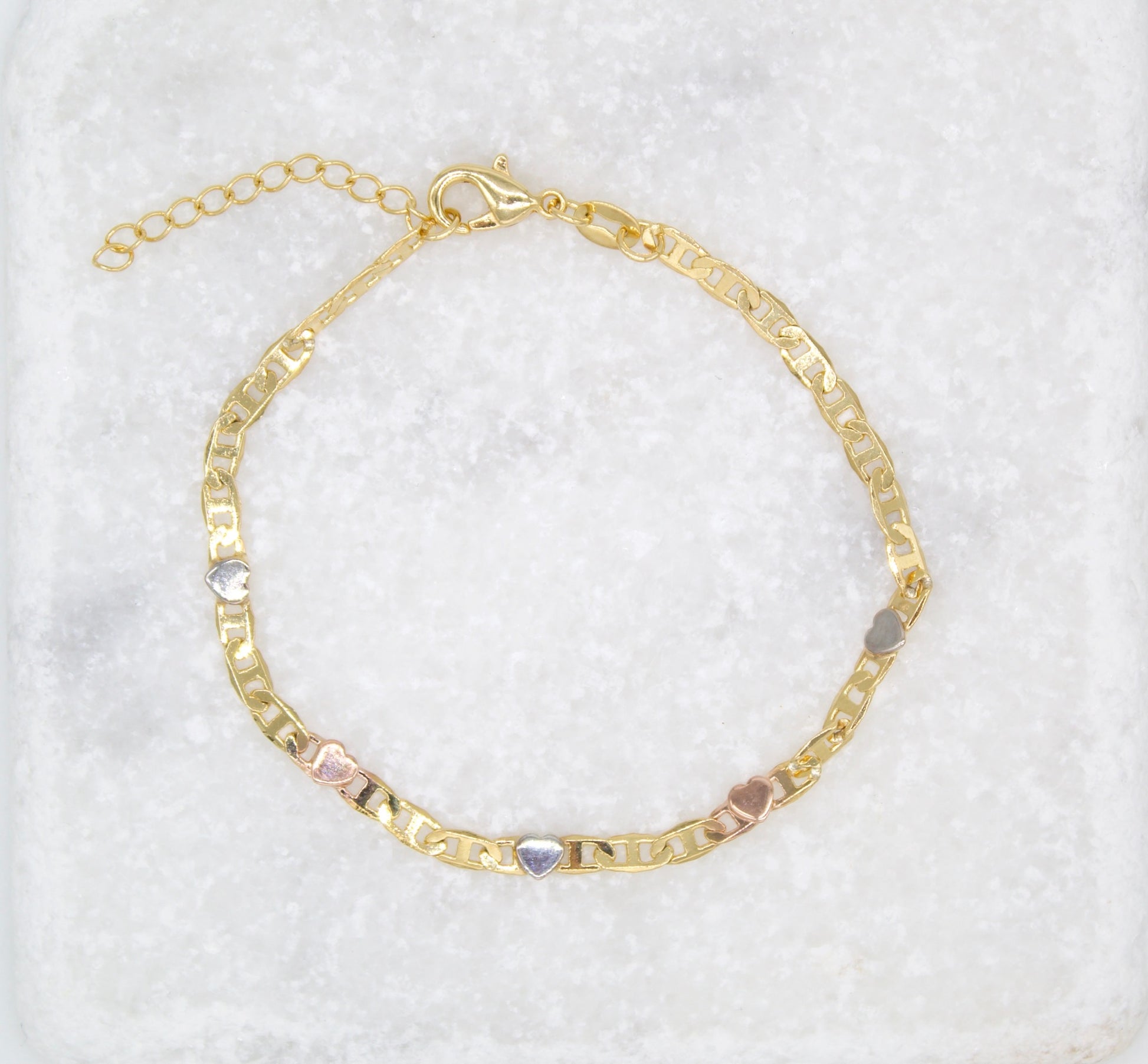 A delicate gold chain bracelet features alternating multi-toned heart charms and rests on a textured white marble surface.