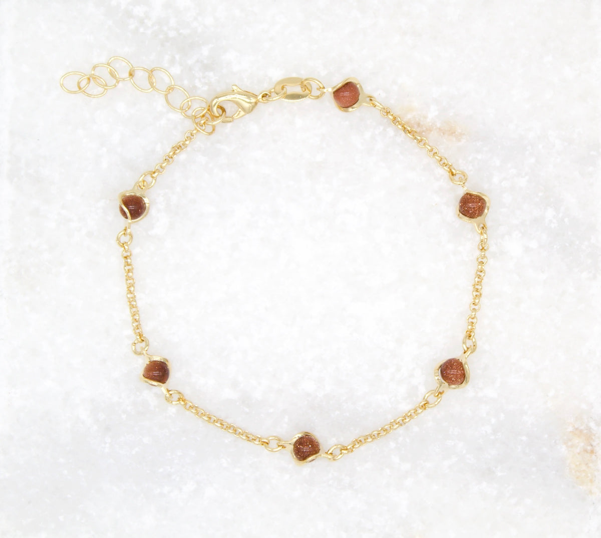 A delicate gold chain bracelet features sparkling brown sunstone beads laid out against a white marble background.