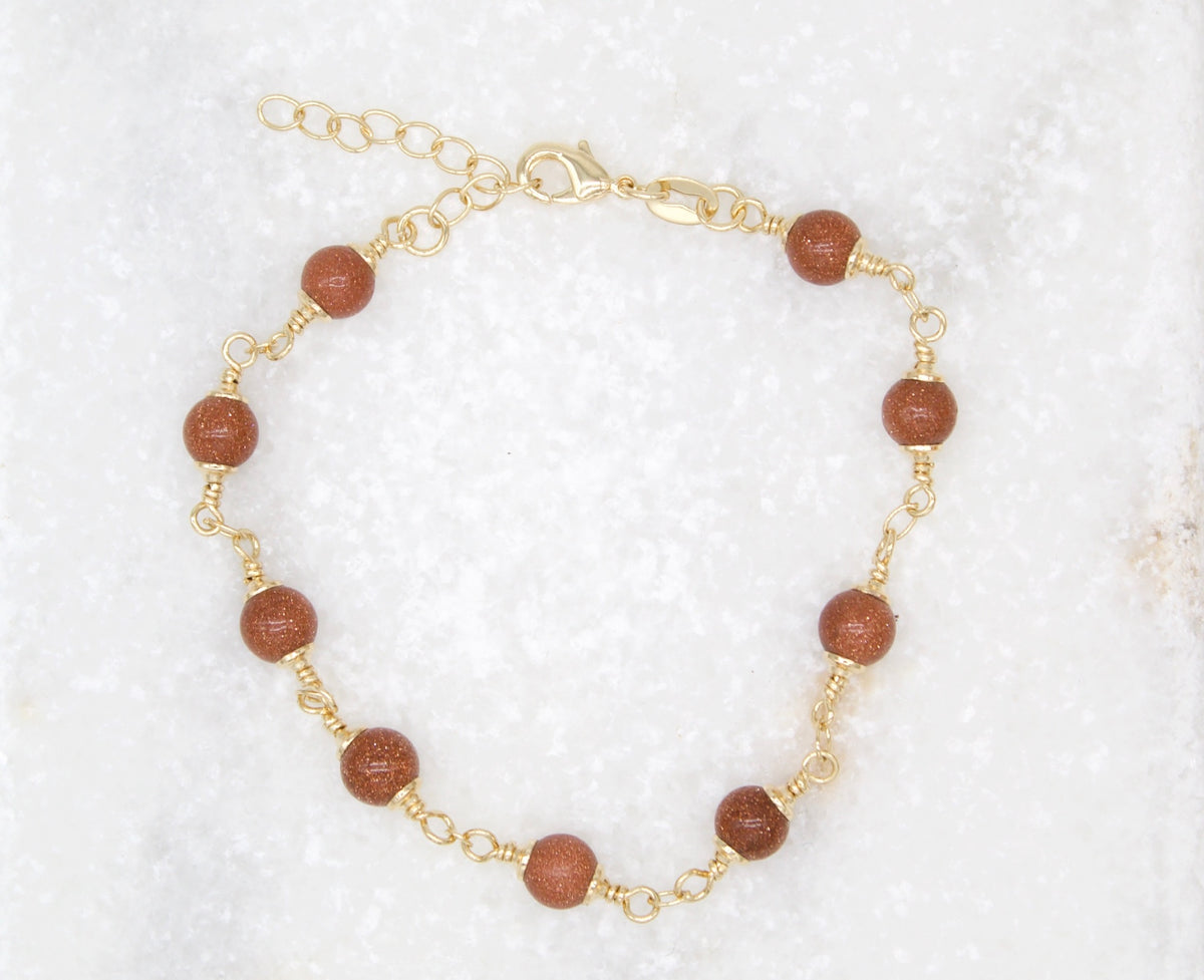 A gold chain bracelet features sparkling brown sandstone gemstone beads arranged in a circle on a white background.