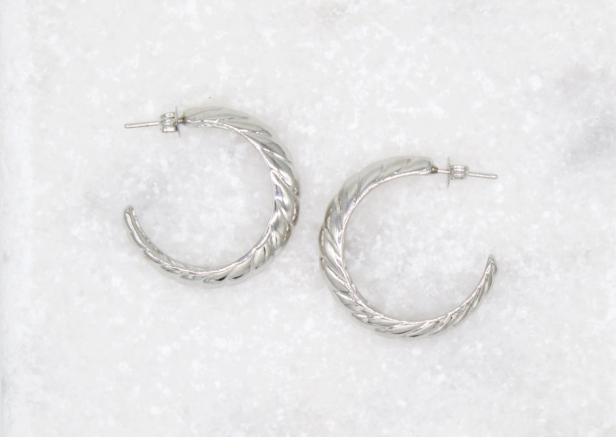 A pair of silver twisted hoop earrings rests on a textured white surface.