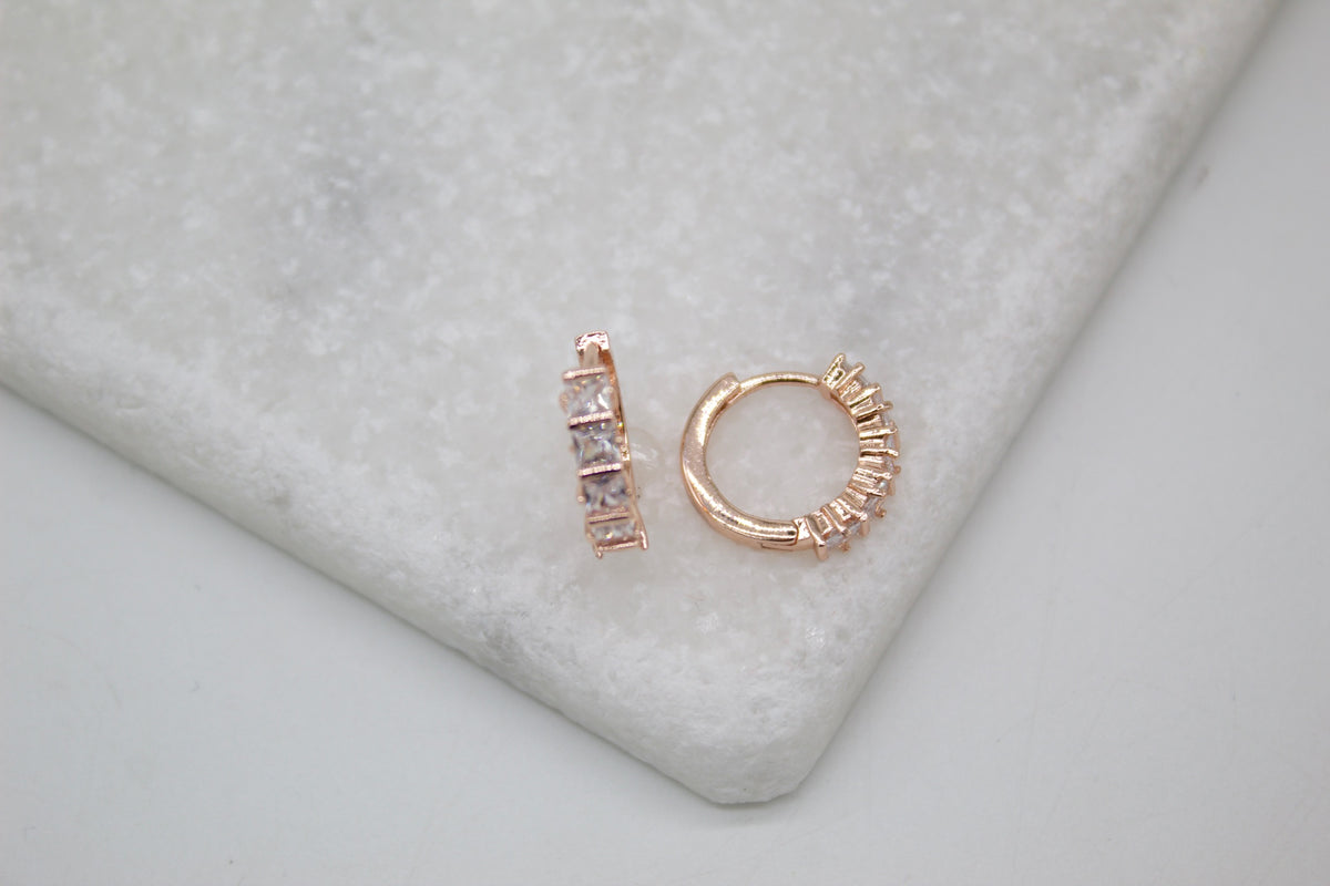 A pair of sparkling rose gold huggie hoop earrings resting on a white marble surface.