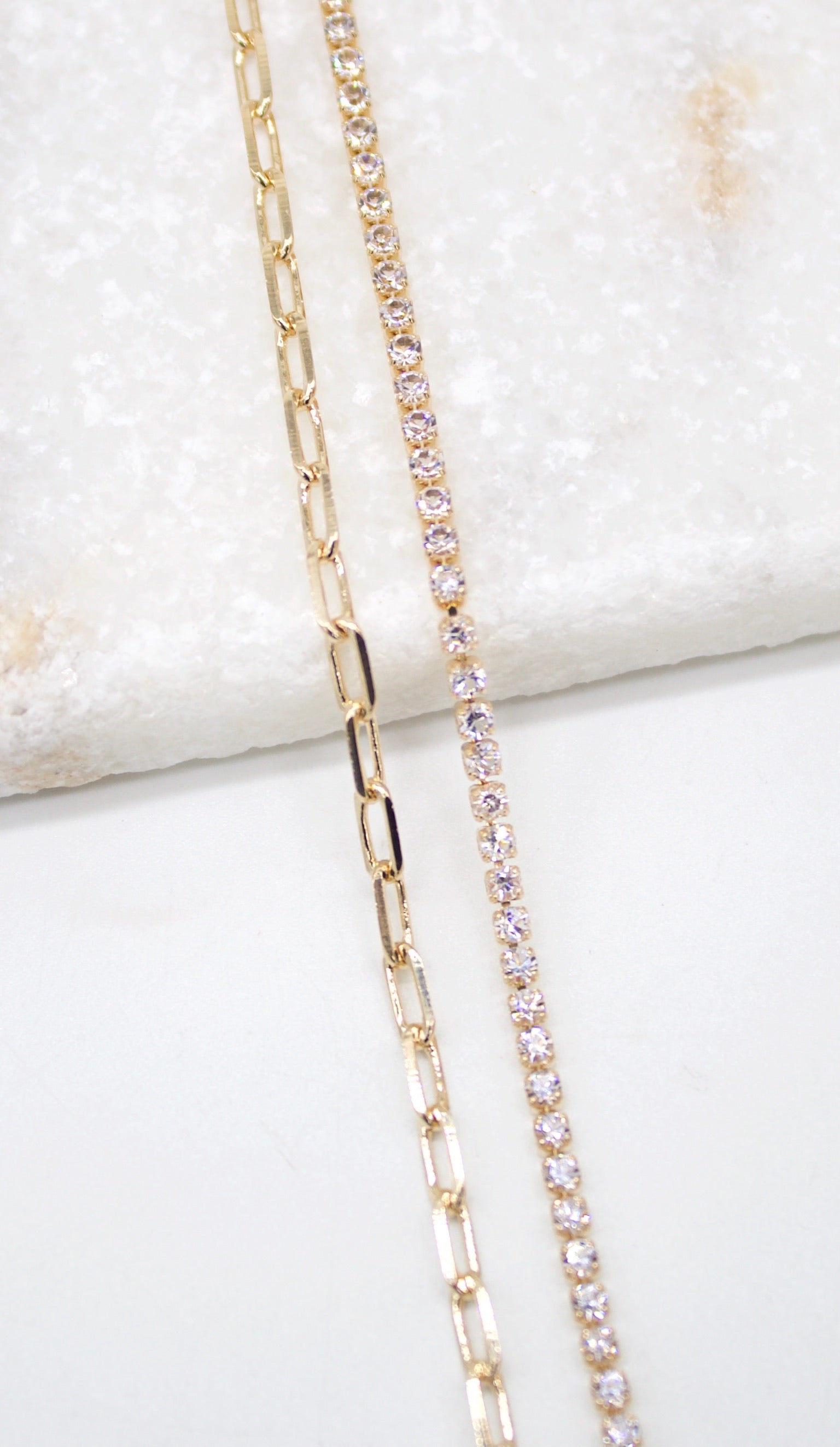 Two delicate gold necklaces, one a paperclip chain and the other a sparkling crystal tennis necklace, rest side by side on a textured white surface.