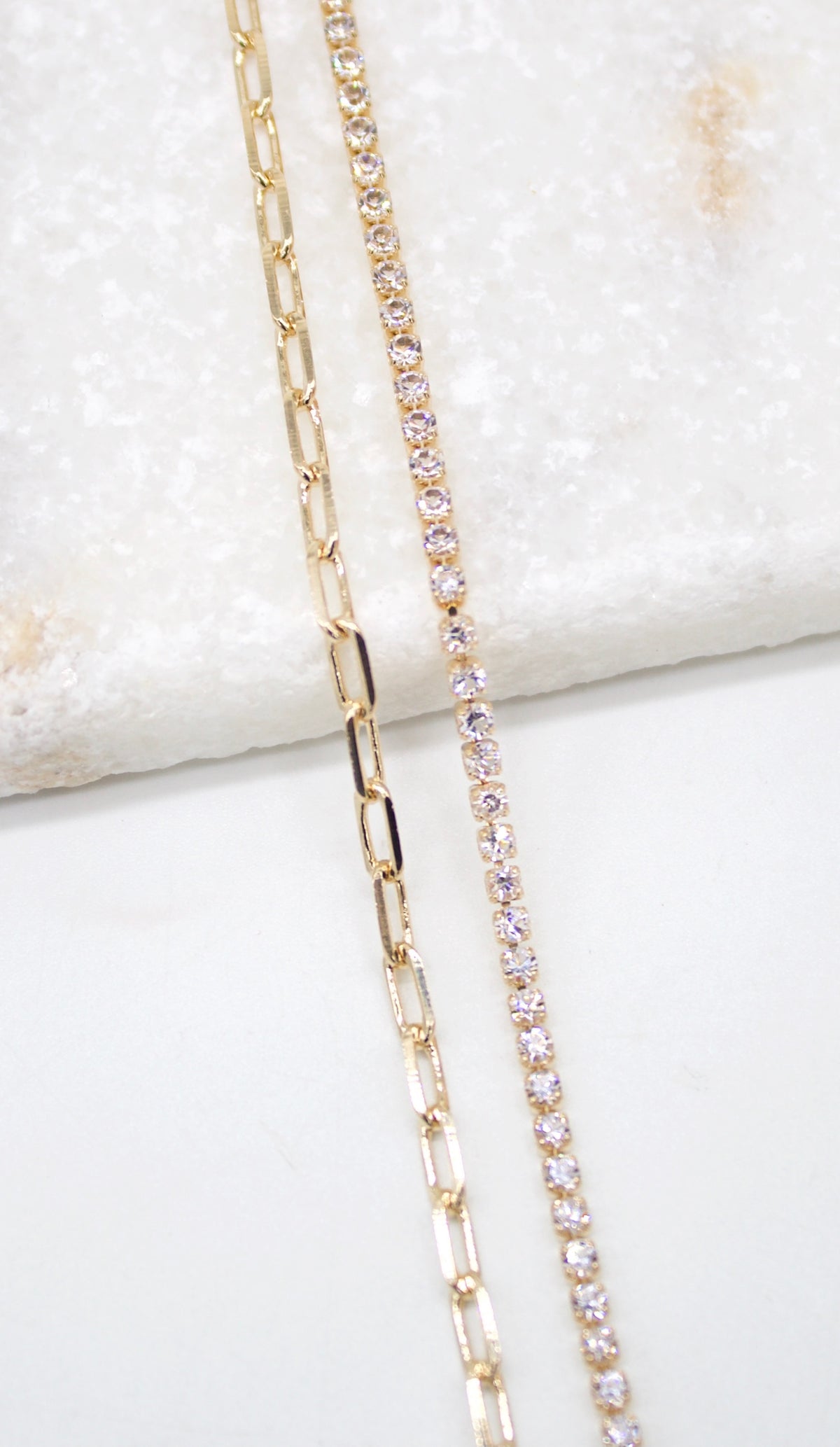 A gold paperclip chain rests beside a sparkling rhinestone tennis bracelet on a white marble surface.