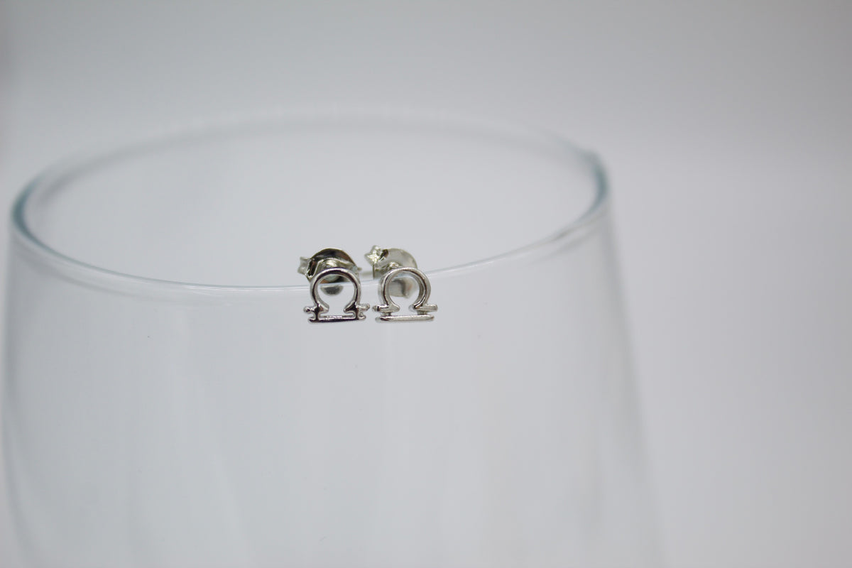 A pair of sterling silver Libra zodiac symbol stud earrings rests on the rim of a clear glass.