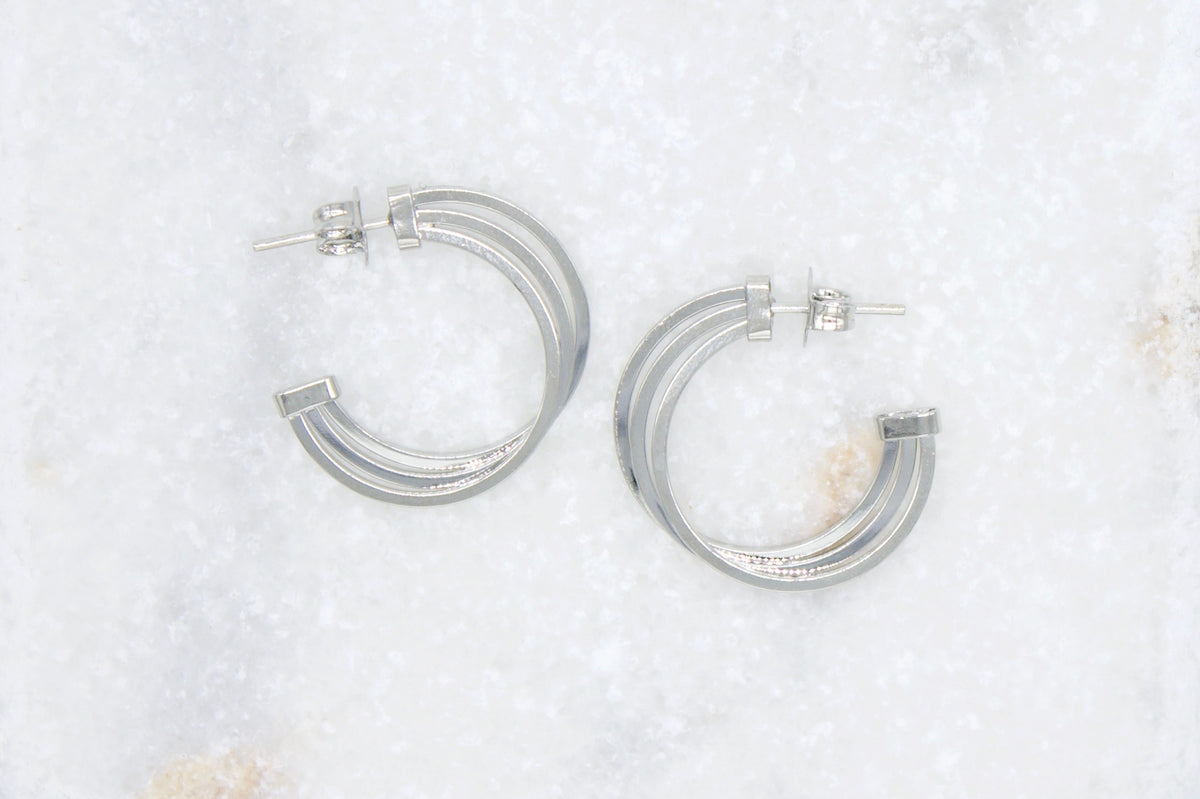 A pair of silver triple hoop earrings rests on a textured white marble surface.