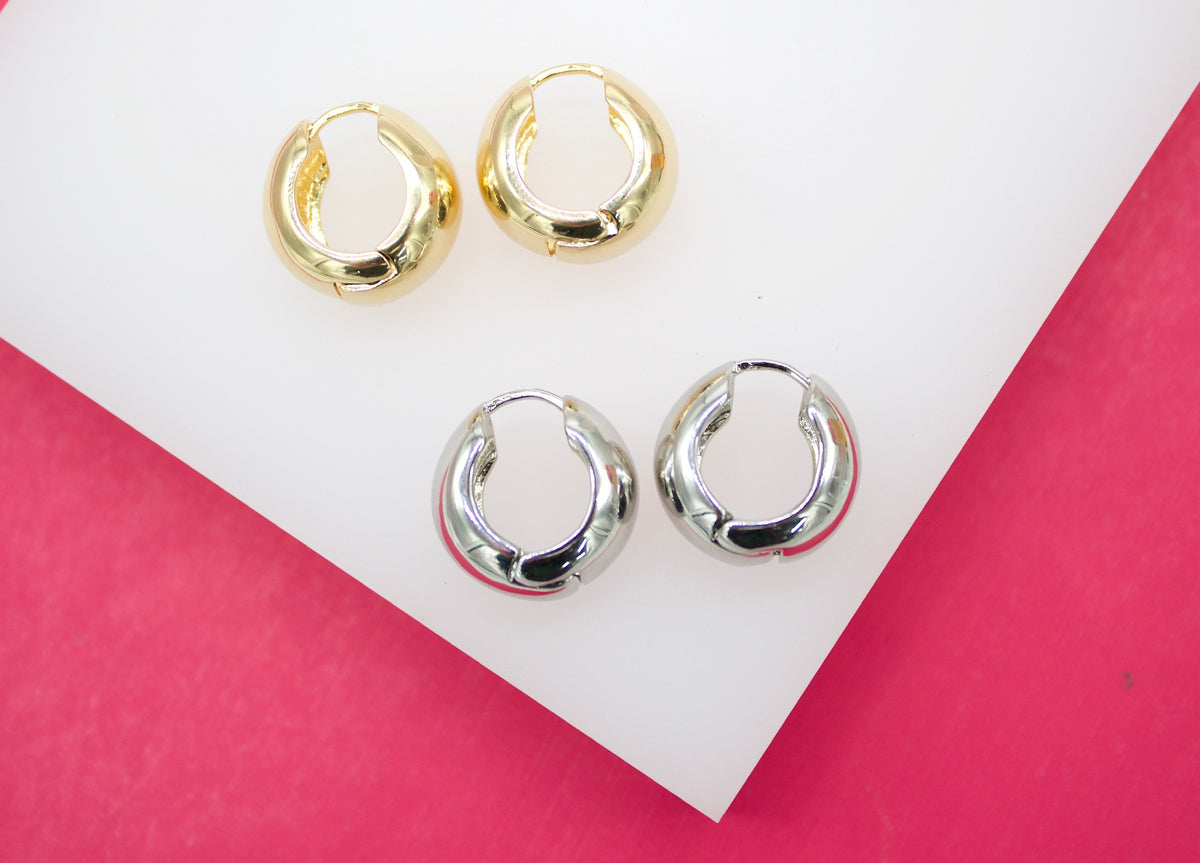 Two pairs of shiny gold and silver small huggie hoop earrings rest on a white display against a vibrant pink background.