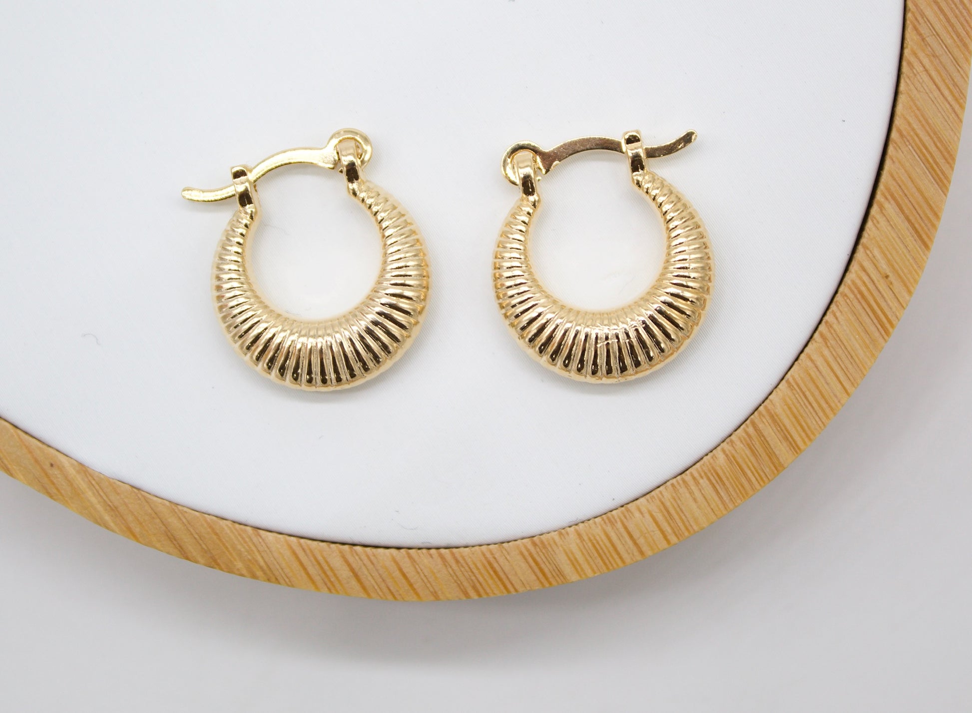 A pair of textured gold hoop earrings rests on a white display tray with a wooden rim.