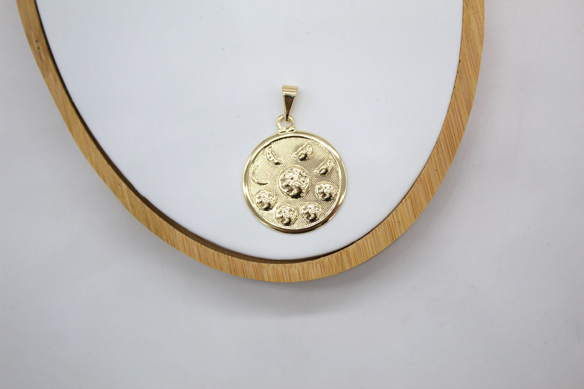 A round gold pendant featuring celestial sun and moon motifs rests on a white display surface with a wooden frame.