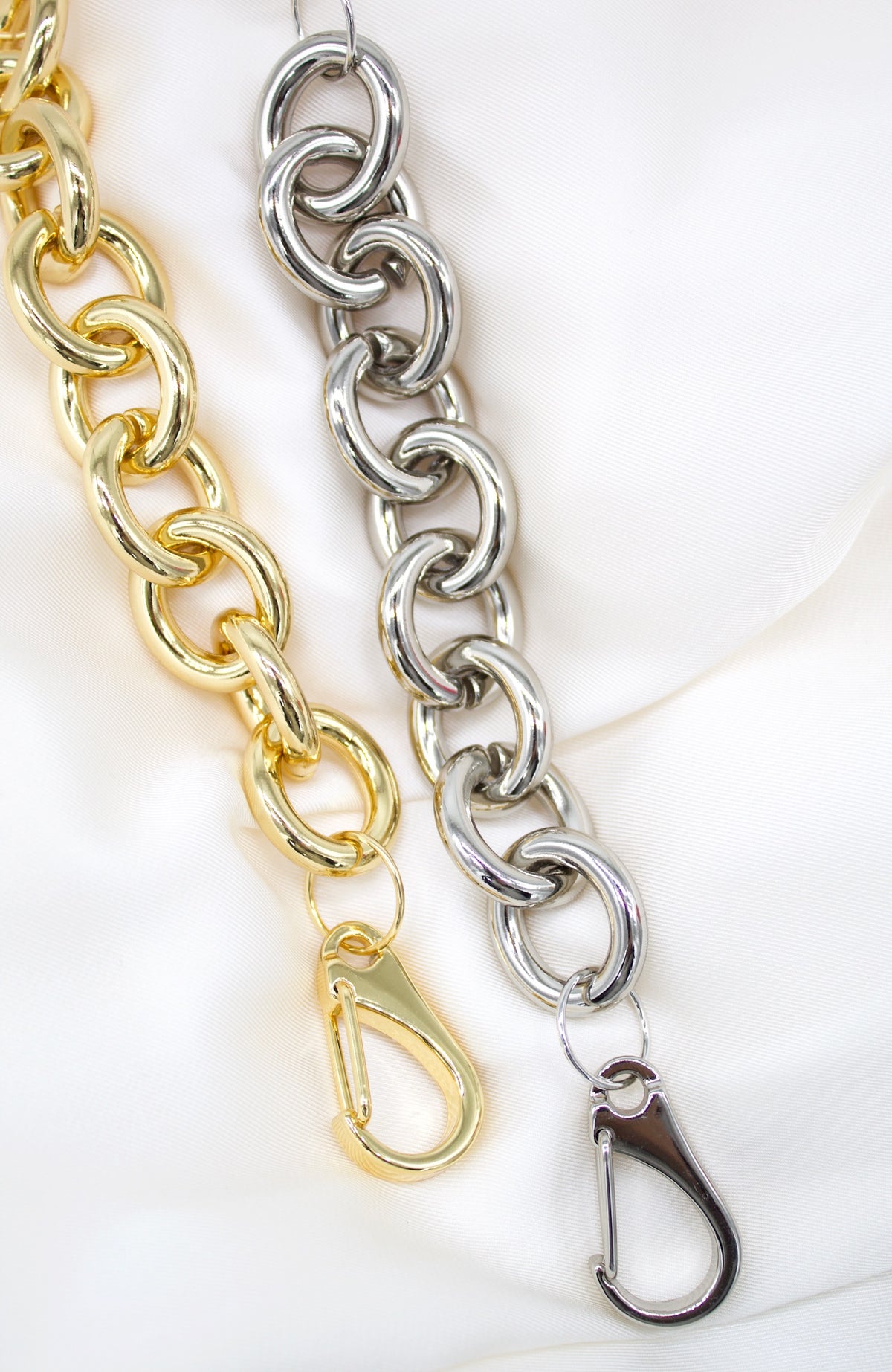 Two chunky link chains, one in gold and one in silver, rest against a white satin background, each featuring a polished lobster claw clasp.