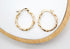 A pair of twisted gold hoop earrings rests neatly on a white display tray with a wooden rim.