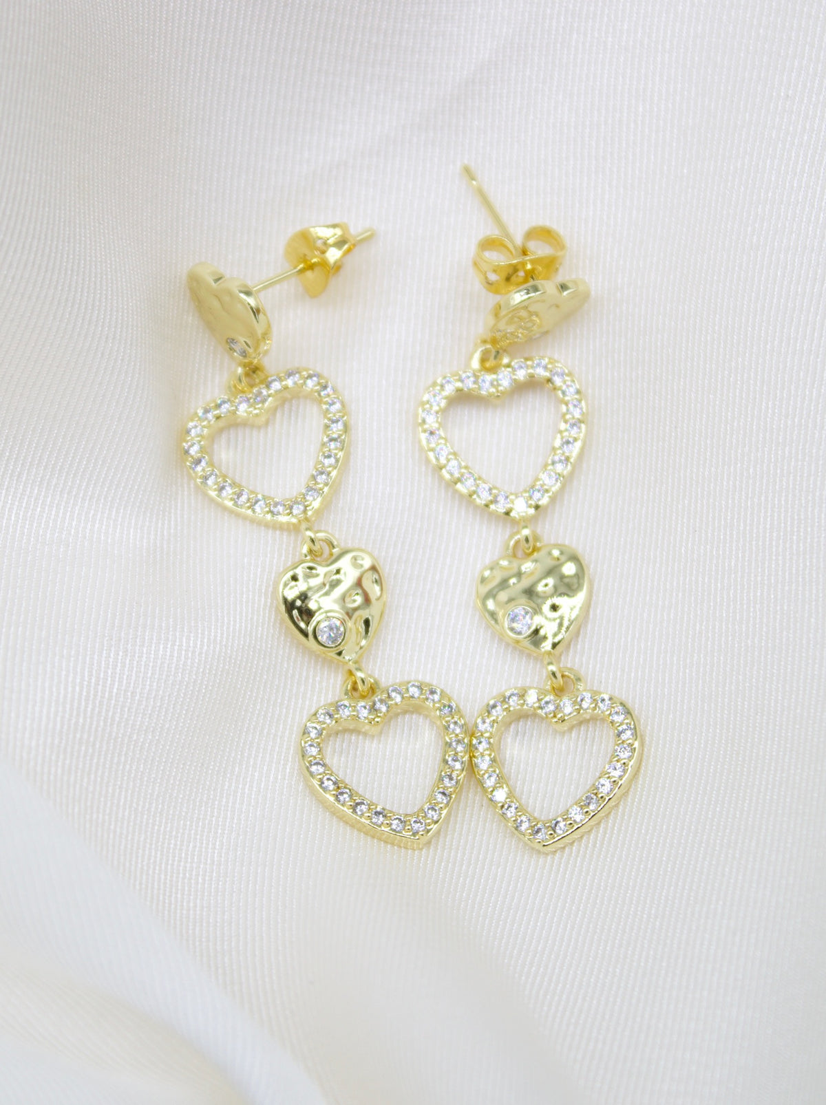 A pair of gold drop earrings featuring alternating pave-set crystal hearts and hammered solid hearts rests on a white satin background.
