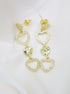 A pair of gold drop earrings featuring alternating pave-set crystal hearts and hammered solid hearts rests on a white satin background.