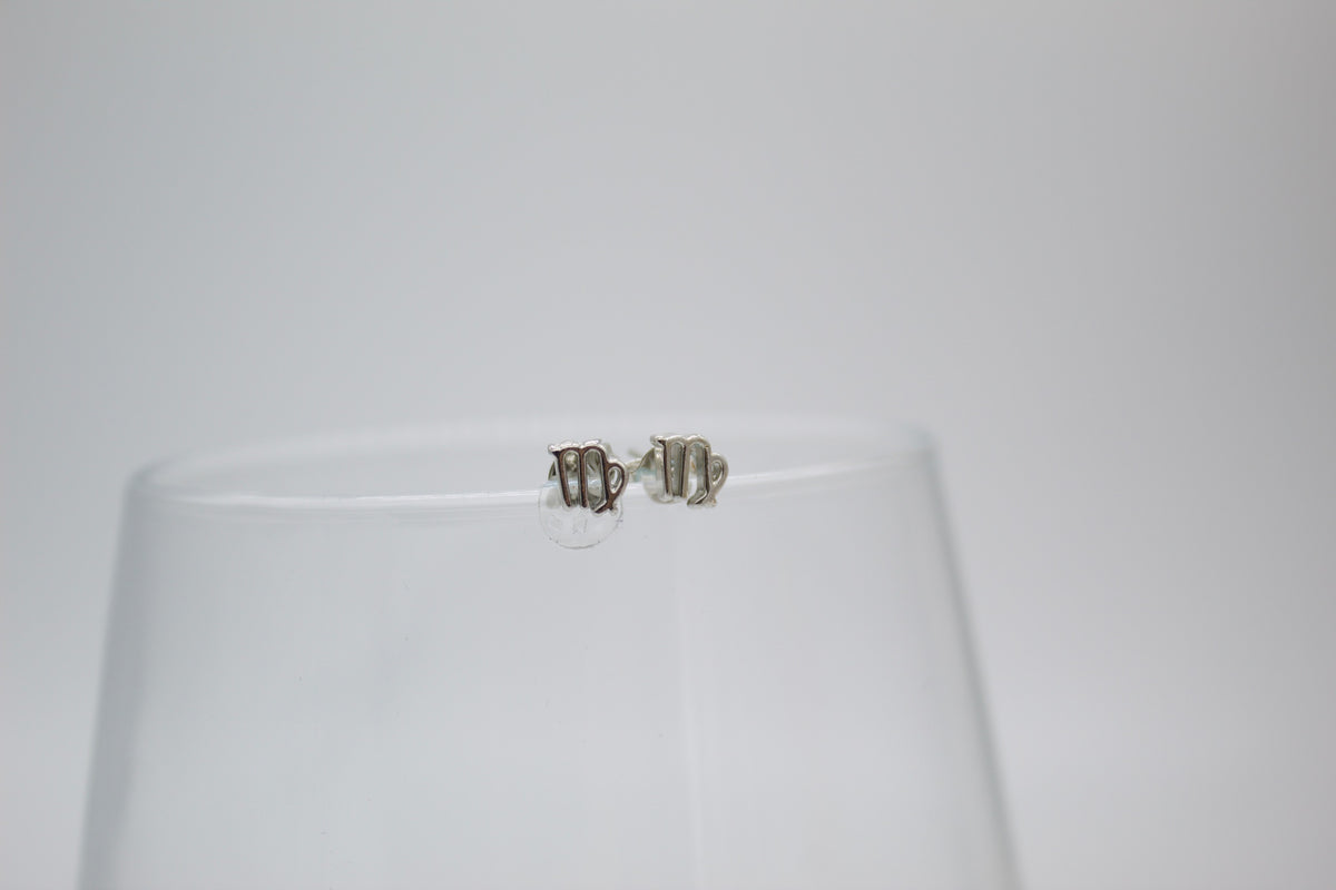A pair of silver Virgo zodiac sign stud earrings rests on the rim of a clear glass.