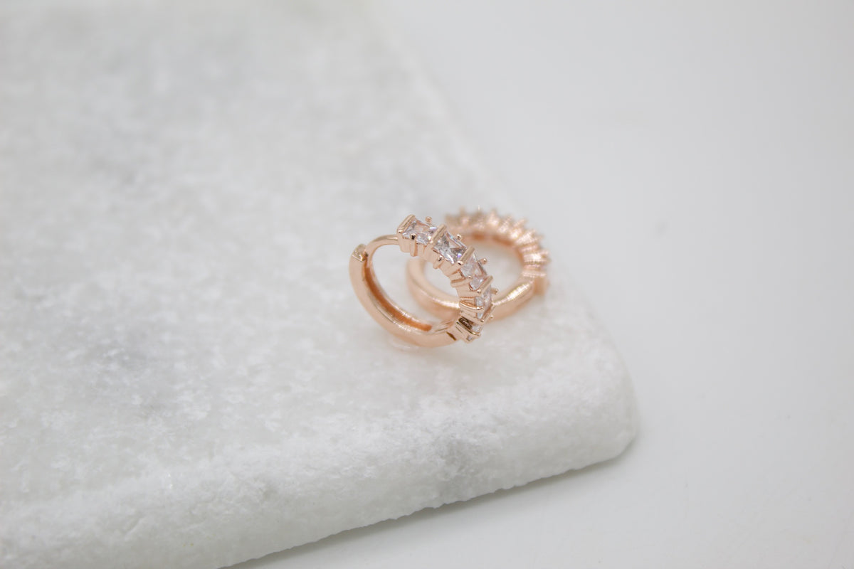 A pair of sparkling rose gold huggie hoop earrings rests on a white marble surface.
