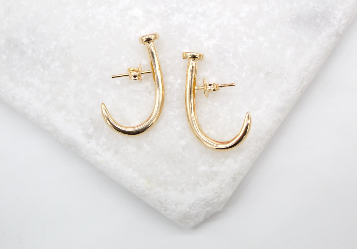 A pair of gold nail-shaped stud earrings rests on a textured white marble surface.