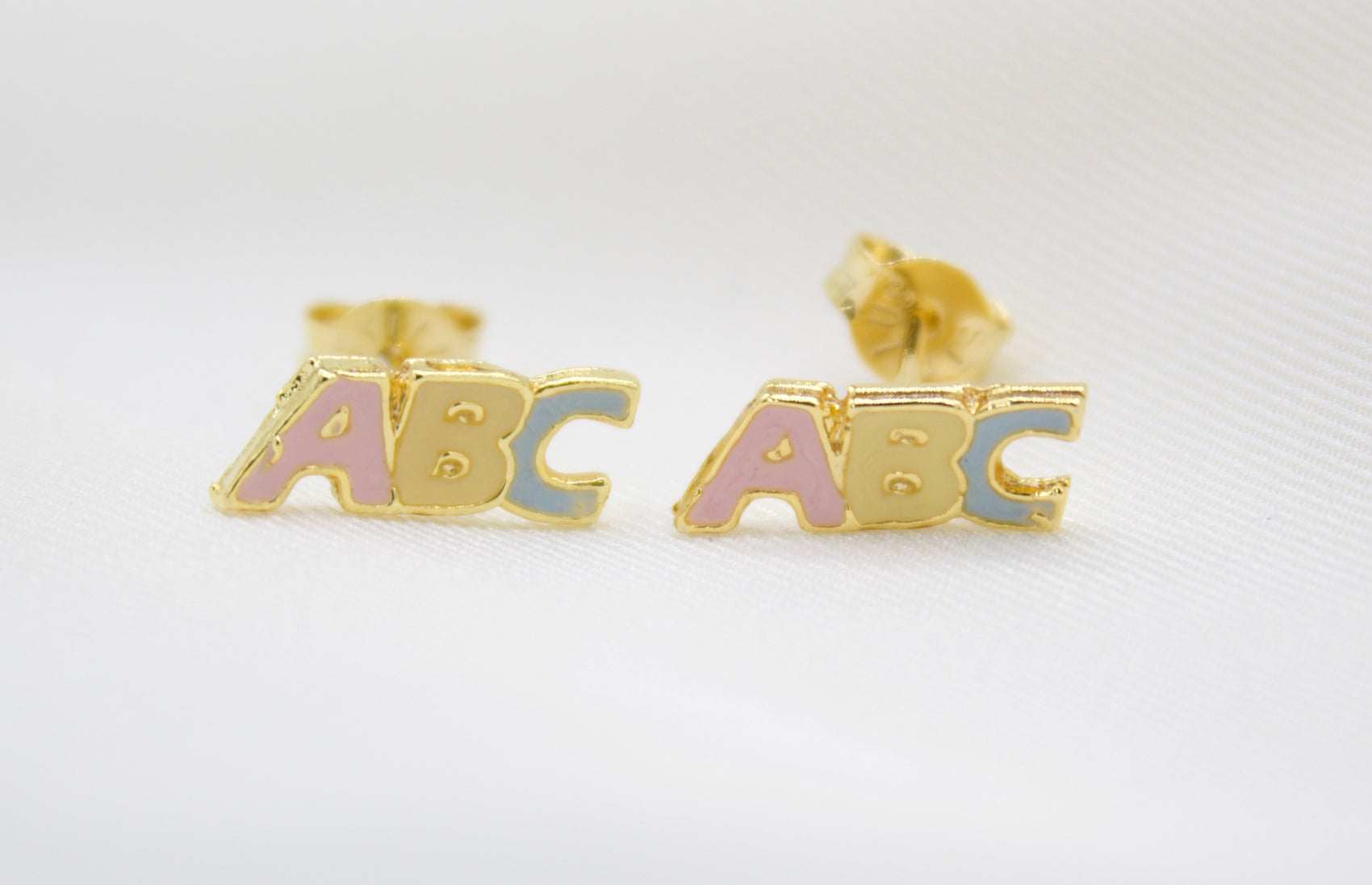 A pair of gold ABC alphabet stud earrings features colorful enamel letters on a white background.
