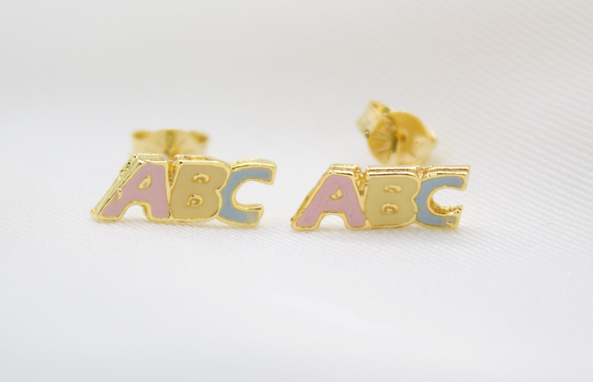 A pair of gold ABC alphabet stud earrings features colorful enamel letters on a white background.