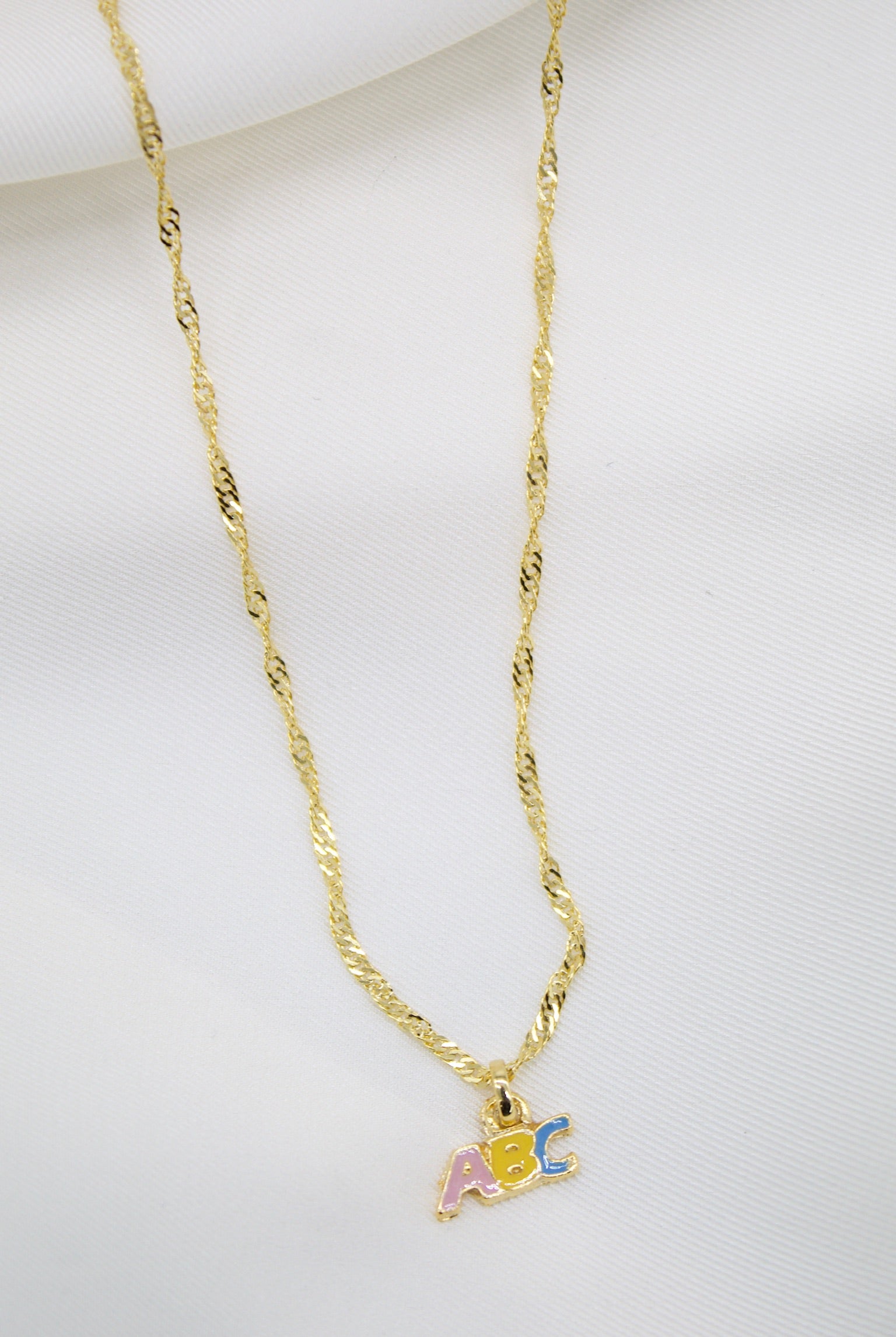 A delicate gold chain necklace displays a colorful ABC pendant resting against a white fabric background.