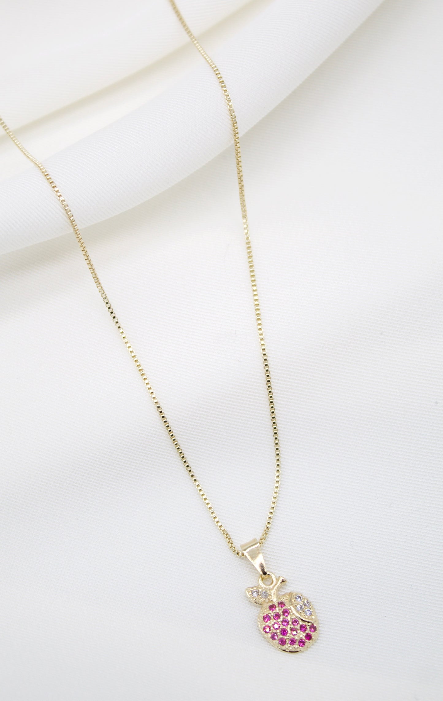 A delicate gold chain necklace drapes against a soft white fabric background, featuring a charming apple-shaped pendant encrusted with sparkling pink and white stones.