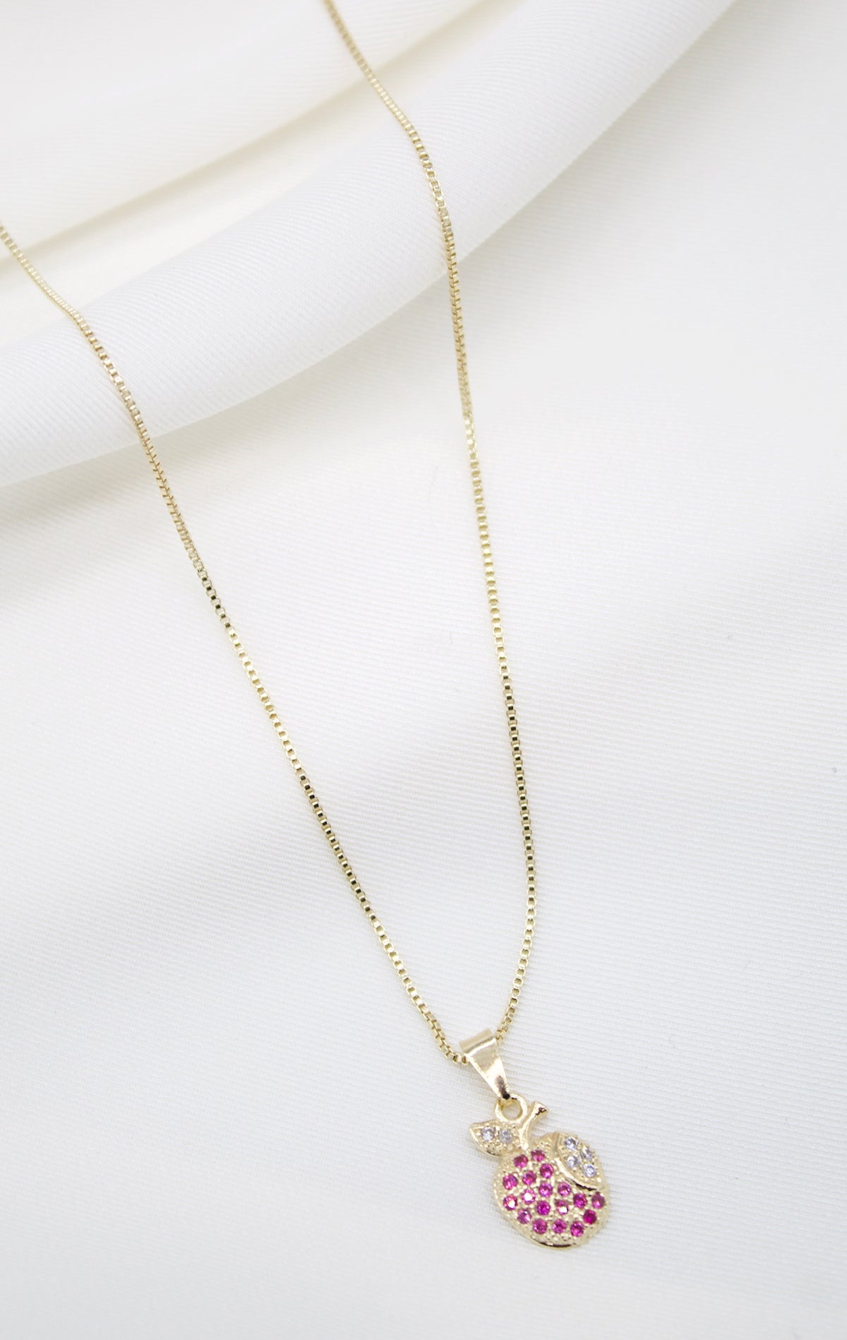 A delicate gold box chain necklace displays a cute apple-shaped pendant encrusted with vibrant pink and white cubic zirconia stones against a soft white fabric background.