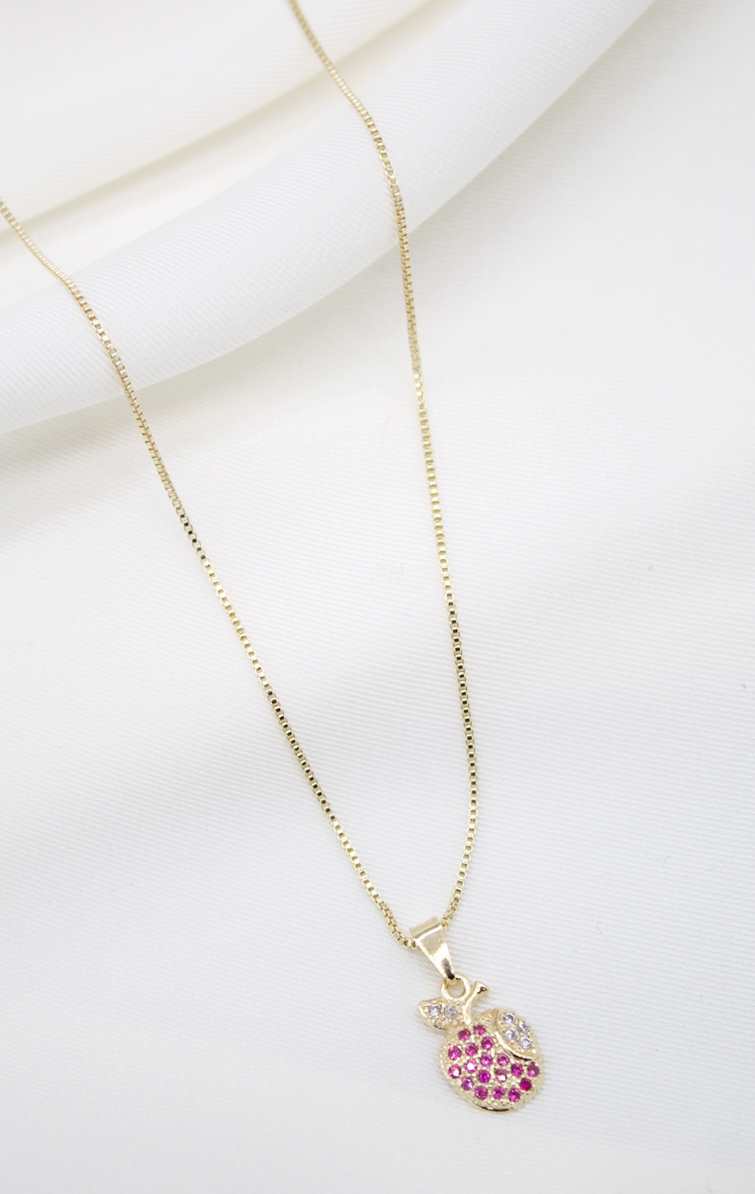 A delicate gold chain necklace drapes against a soft white fabric background, featuring a charming apple-shaped pendant encrusted with sparkling pink and white stones.