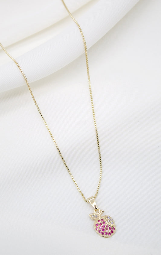 A delicate gold chain necklace drapes against a soft white fabric background, featuring a charming apple-shaped pendant encrusted with sparkling pink and white stones.