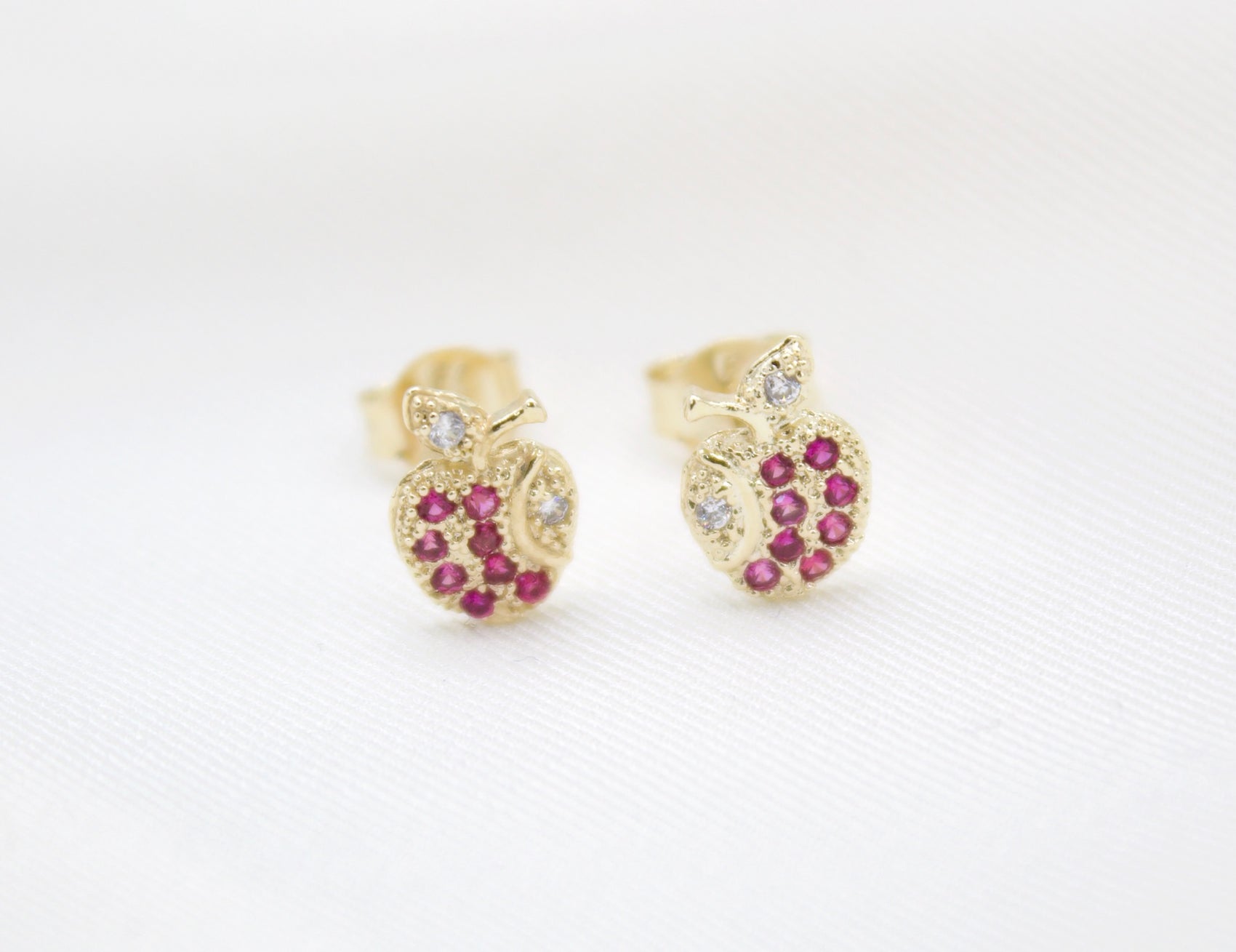 A pair of gold apple-shaped stud earrings sparkle with ruby red gemstones and small white diamonds on a white fabric background.