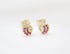 A pair of gold apple-shaped stud earrings sparkle with ruby red gemstones and small white diamonds on a white fabric background.