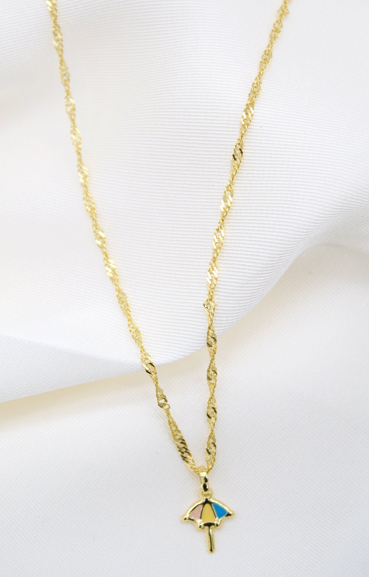 A delicate gold chain necklace with a colorful umbrella pendant rests on a soft white fabric background.