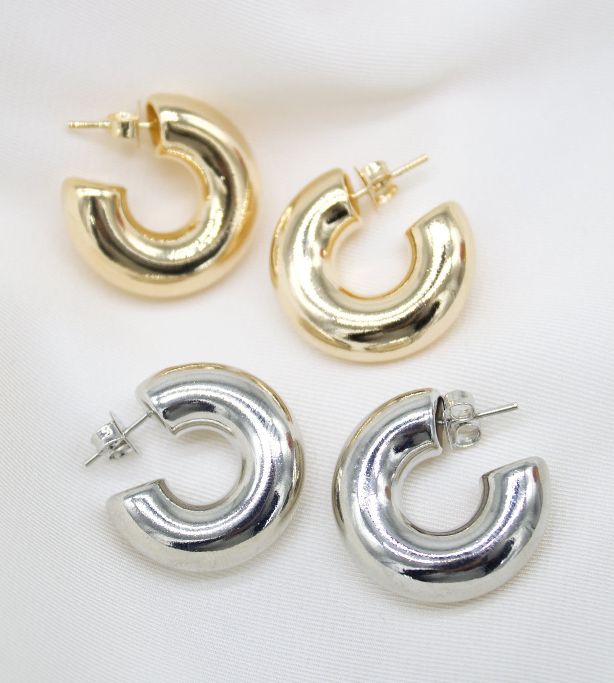 Two pairs of chunky hoop earrings in gold and silver rest neatly on a white fabric background.