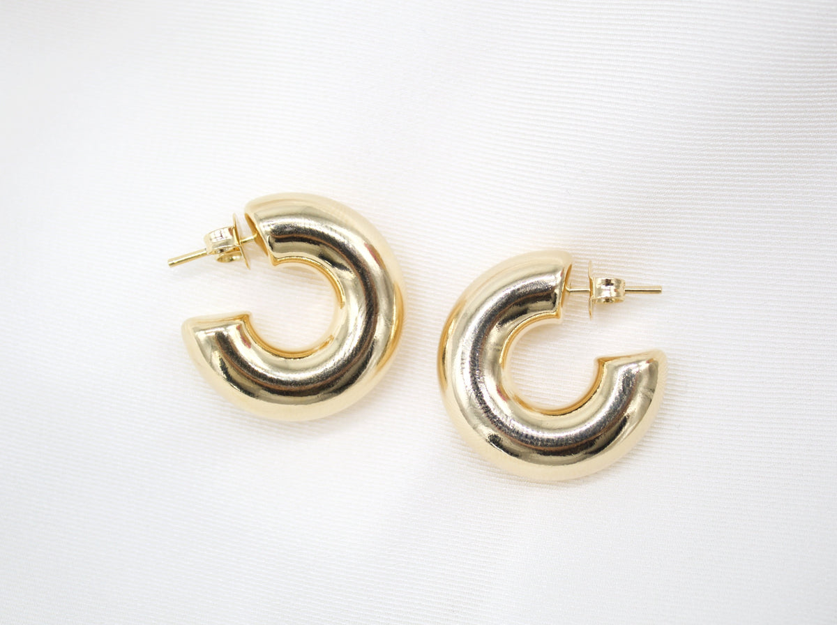 A pair of chunky gold hoop earrings rests on a textured white silk background.