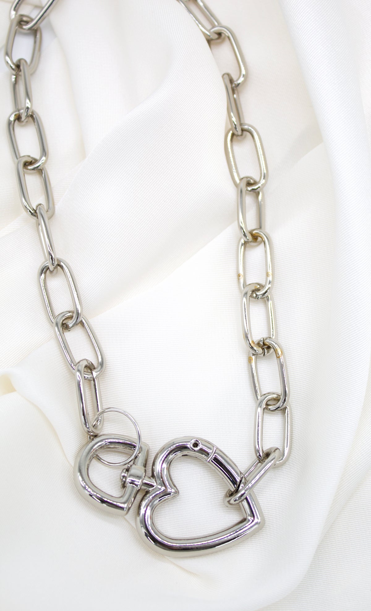 A chunky silver link necklace rests on a white satin background, featuring an interlocking heart clasp at the center.