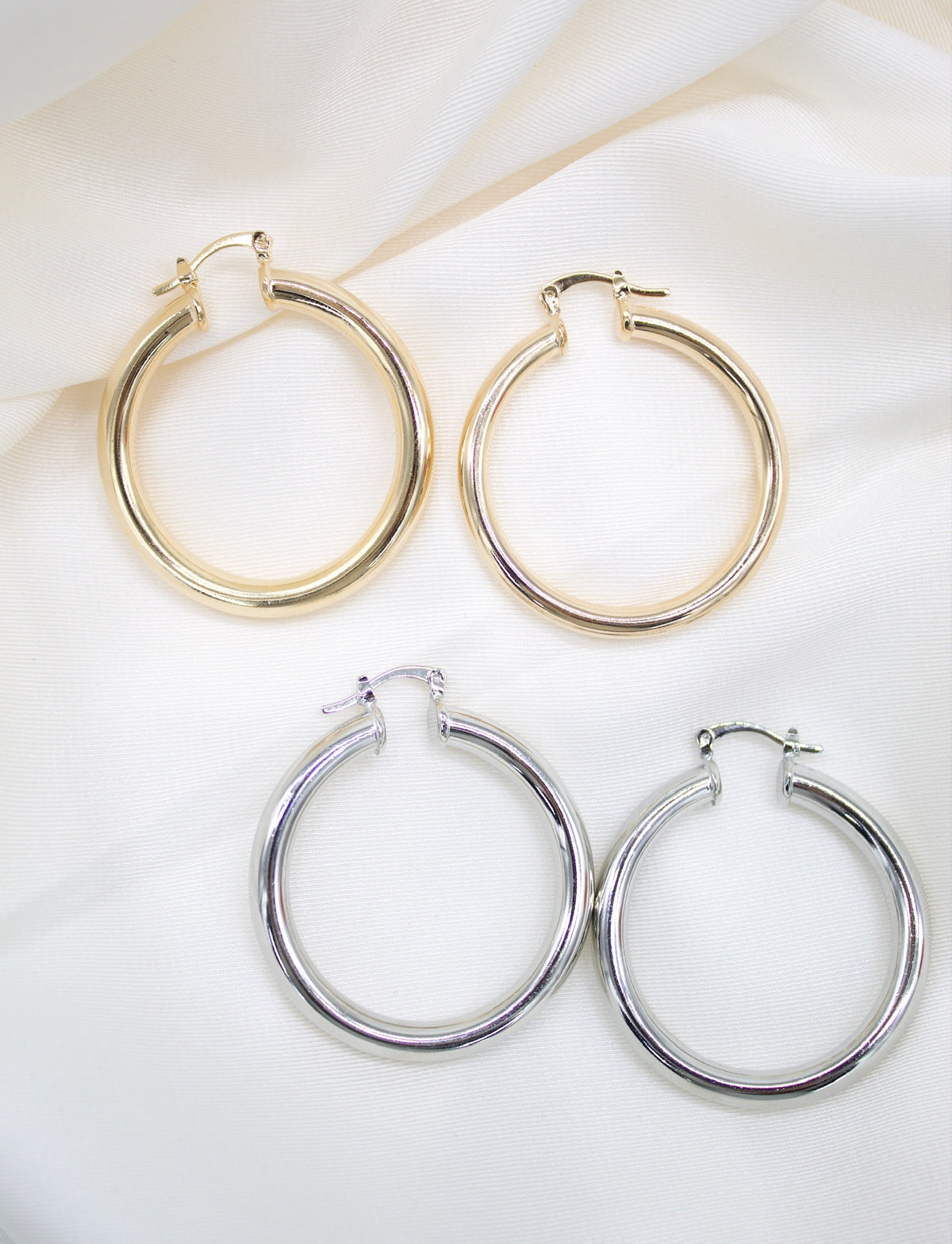 Two pairs of polished gold and silver hoop earrings rest on a white silk background.