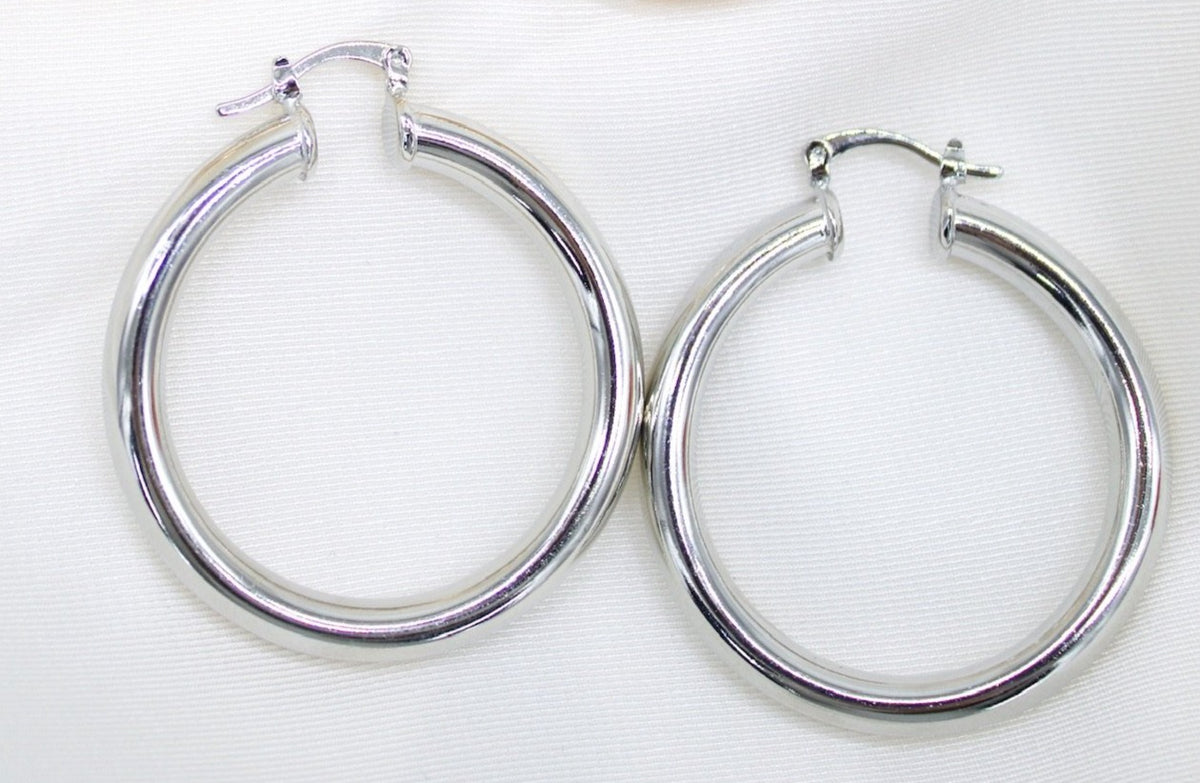 A pair of sleek silver hoop earrings rests on a soft white fabric background.