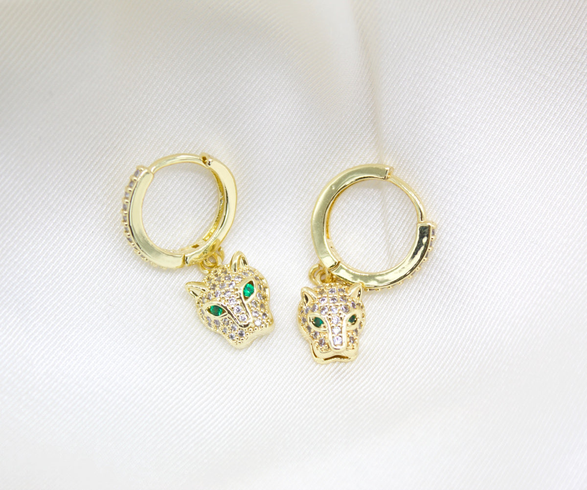 A pair of gold hoop earrings featuring crystal-pave panther charms with vibrant green eyes rests on a soft white satin background.