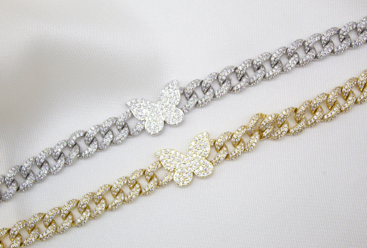 Two iced-out Cuban link bracelets featuring pave-set diamond butterflies rest diagonally on a white silk background.