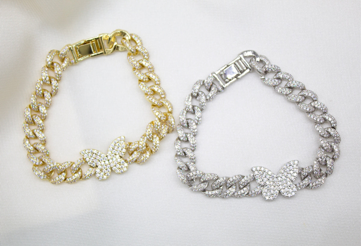 Two icy cuban link chain bracelets adorned with dazzling butterfly charms rest side by side on a satin background, showcasing one in gleaming gold and the other in radiant silver.