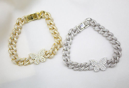Two icy cuban link chain bracelets adorned with dazzling butterfly charms rest side by side on a satin background, showcasing one in gleaming gold and the other in radiant silver.