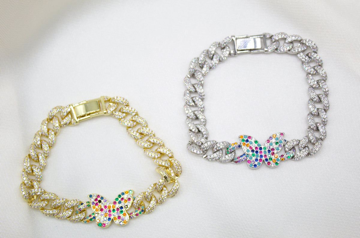 A gold and a silver cuban link butterfly bracelet rest side-by-side on a white satin background.